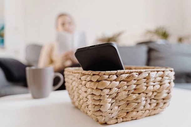 Woman sitting on a couch reading a book while her phone rests in a basket in the foreground.