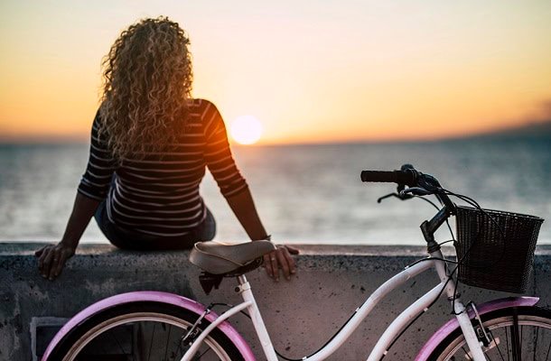 Woman looking at a sunset at the beach with her bike leaning at concrete in the foreground.