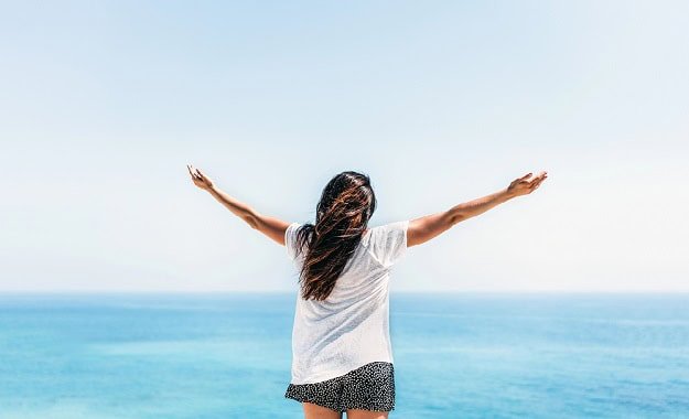 Woman standing in front of the ocean in summer with her arms stretched out.