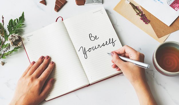 A woman writing Be Yourself in a notebook.