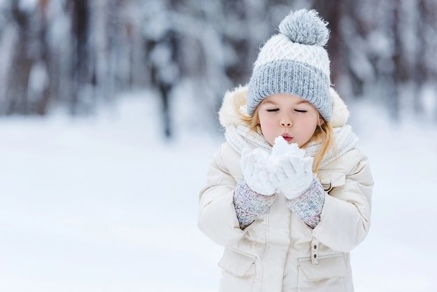 A small girl blowing at snow in her hands while standing in a forest in winter.