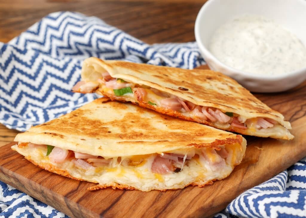 Ham-and-Cheese-Quesadilla-BFK-6-1024x731.jpg A ham and cheese quesadilla has been sliced and is ready to eat, served on a wooden board with a side of dip!