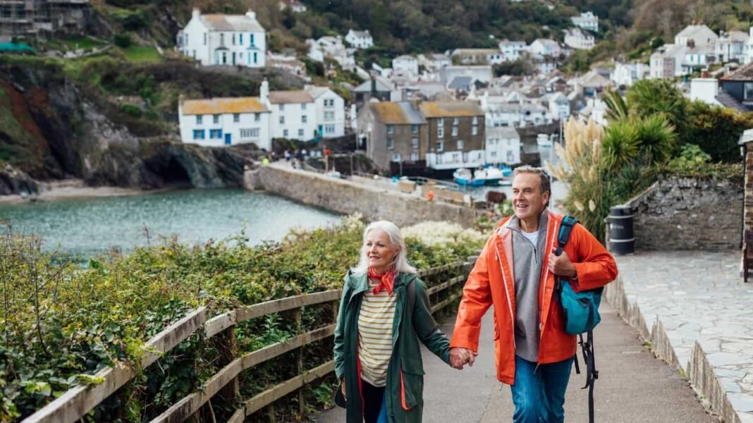 ISA or SIPP? List one advantage and one disadvantage of A senior man and his wife holding hands walking up a hill on a footpath looking away from the camera at the view. The fishing village of Polperro is behind them.