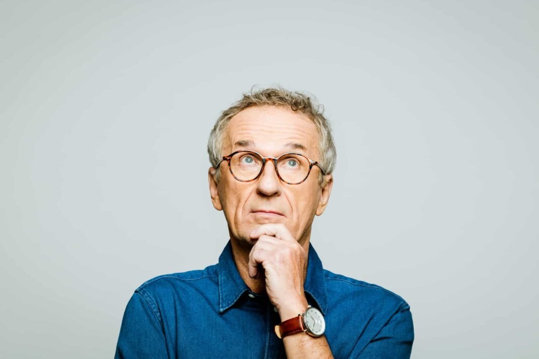 This FTSE 250 stock crashed 18% today! Is it too Portrait of elderly man wearing white denim shirt and glasses looking up with hand on chin. Thoughtful senior entrepreneur, studio shot against grey background.