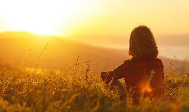 A woman sitting in a field and looking at a sunrise.