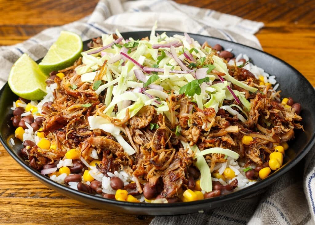 Carnitas-Bowl-BFK-2-1024x731.jpg A horizontally aligned image of a pork carnitas bowl piled high with cabbage, rice, beans, corn, limes, and avocadoes, ready to eat!