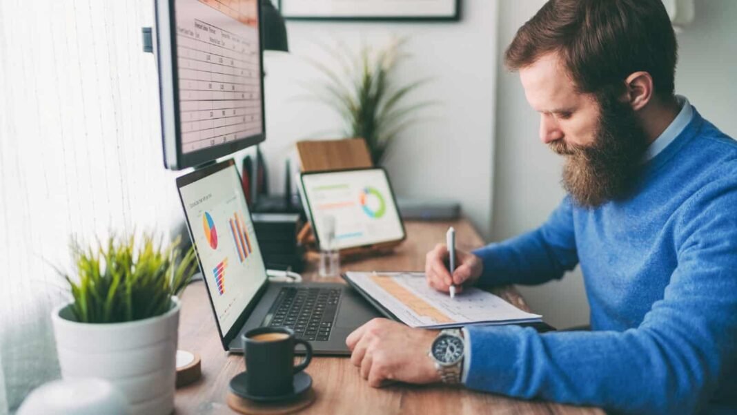 Bearded man writing on notepad in front of computer