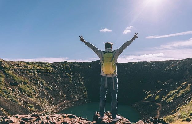 A man at the top of a hike looking out over a water filled crater. It