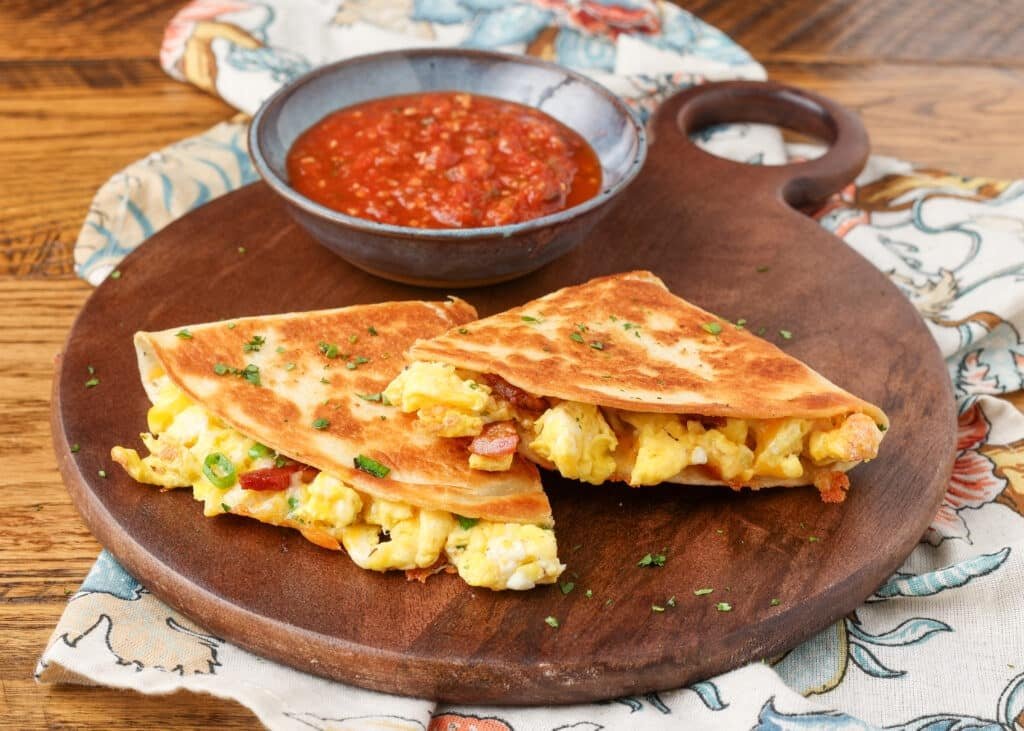 Two wedges of breakfast quesadilla on a wooden platter with a serving of salsa.