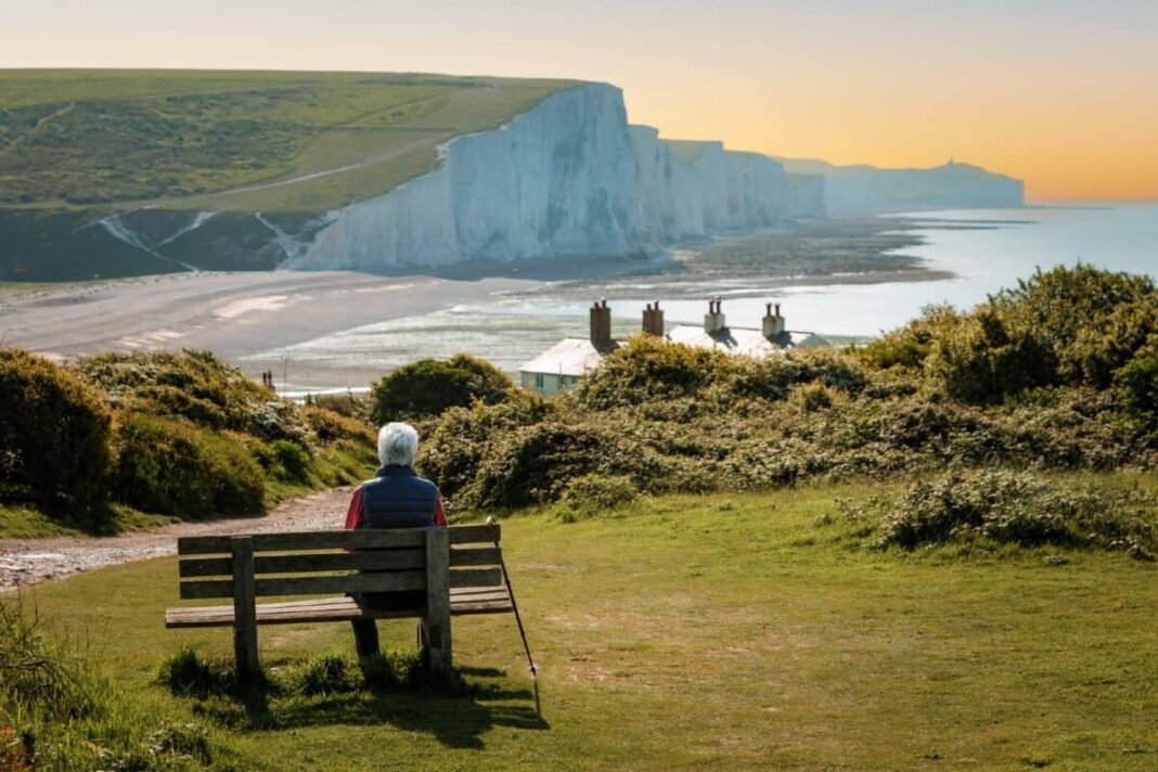 How to earn tax-free ISA income worth double your state Rear view image depicting a senior man in his 70s sitting on a bench leading down to the iconic Seven Sisters cliffs on the coastline of East Sussex, UK. The man is wearing casual clothing - blue denim jeans, a red checked shirt, navy blue gilet. The man is having a rest from hiking and his hiking pole is leaning up against the bench.